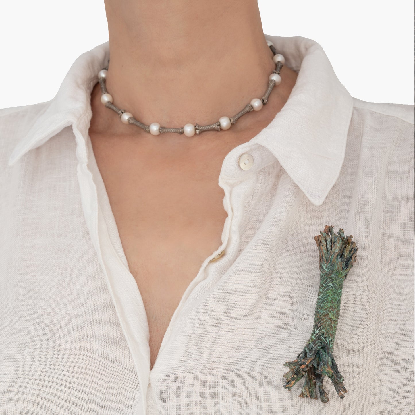 Person wearing a white shirt with an undone top button, a necklace with spherical beads, and a multicolored fabric brooch on the left side of the collar, enhanced by the unique Rope brooch inspired by the San Telmo ship - in memoriam- crafted using an electroforming technique.