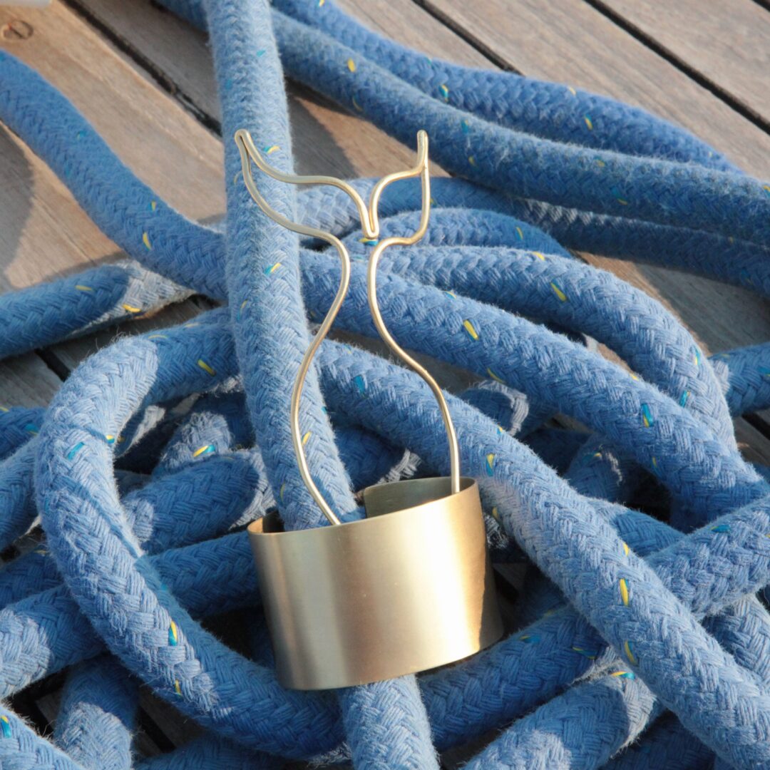 A 24k gold plated brass Whale Tail Cuff Bracelet, inspired by sailor tattoos and handmade by Angela Lago, sits among coiled blue ropes on a wooden surface.